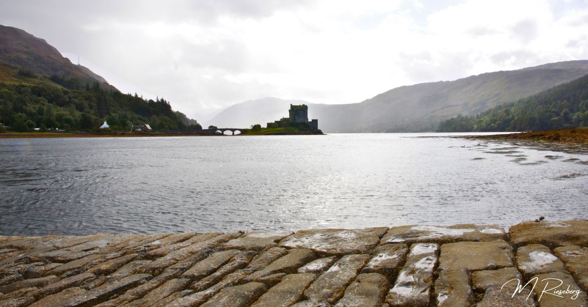 Eilean Donan castle in the Highlands, Scotland in the distance of the image. A big, gray castle surrounded by hills and mountains and then the lake. This photo was taken from a viewpoint further away, across the lake, on a boat ramp made of old stones.