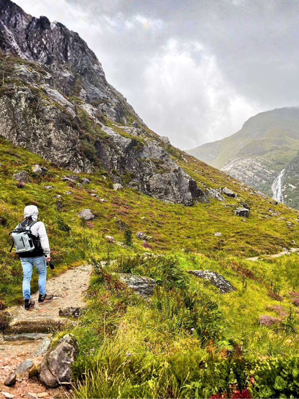 Matze walking a dirt path in the middle of a stunning landscape with the most vivid greens and pops of purple and red flowers and a rocky hill on the left. In the distance you can see more rolling hills and the giant Steall Waterfall in the Highlands in Scotland.