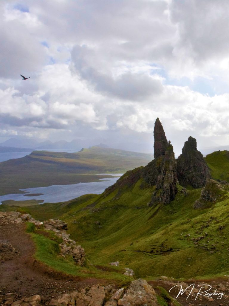 Image of the Old Man of Storr, an imposing rock formation of jagged, tall stacks that rise up, out of grassy, rolling hills into a cloudy sky. A bird flies by in the upper left corner. The ocean is visible in the very far distance and a small lake is visible in the nearer distance. There is a small trial visible, indicating the hike to get to the high up viewpoint. Photo by Matze of Oso Osito.