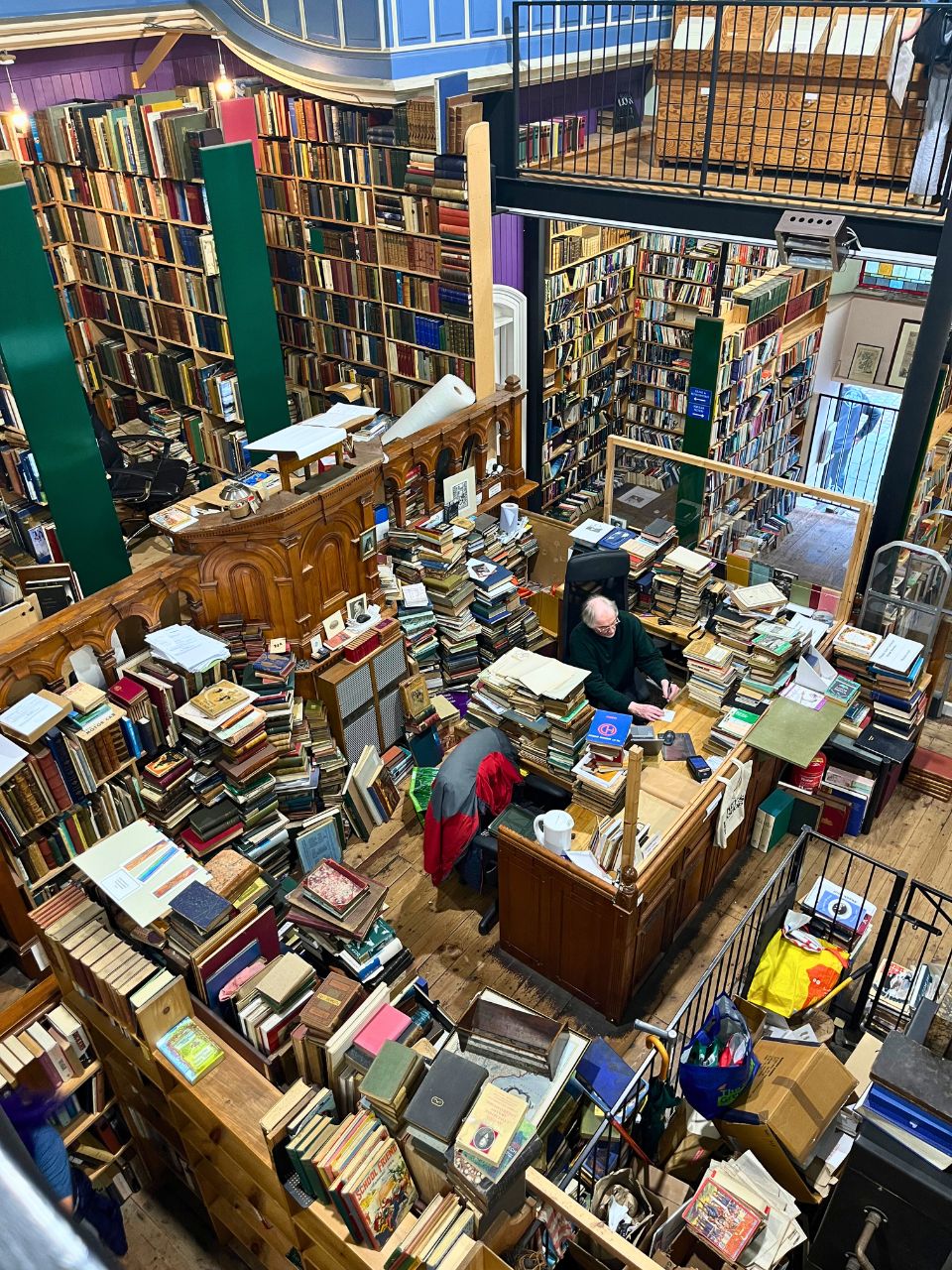 Taken looking down from the second level of a jam packed bookshop where every, single surface is covered with stacks of books. There must be over a thousand books in this image, if not many more. From ancient texts that are falling apart to bright kids books to notebooks and so much more. In the center, on the ground floor, in the distance, is a desk that is also covered in books and there sits a man - the bookshops owner.