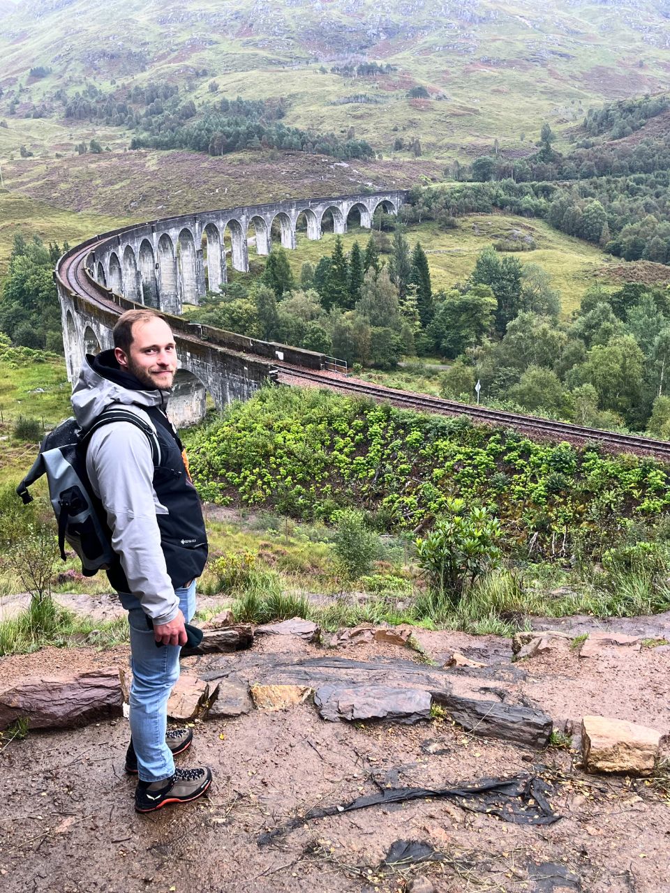 Image of Matze standing on a hill in front of the Glenfinnan Viaduct in Scotland. The viaduct stretches in a half moon shape and is surrounded by tons of lush, green landscape