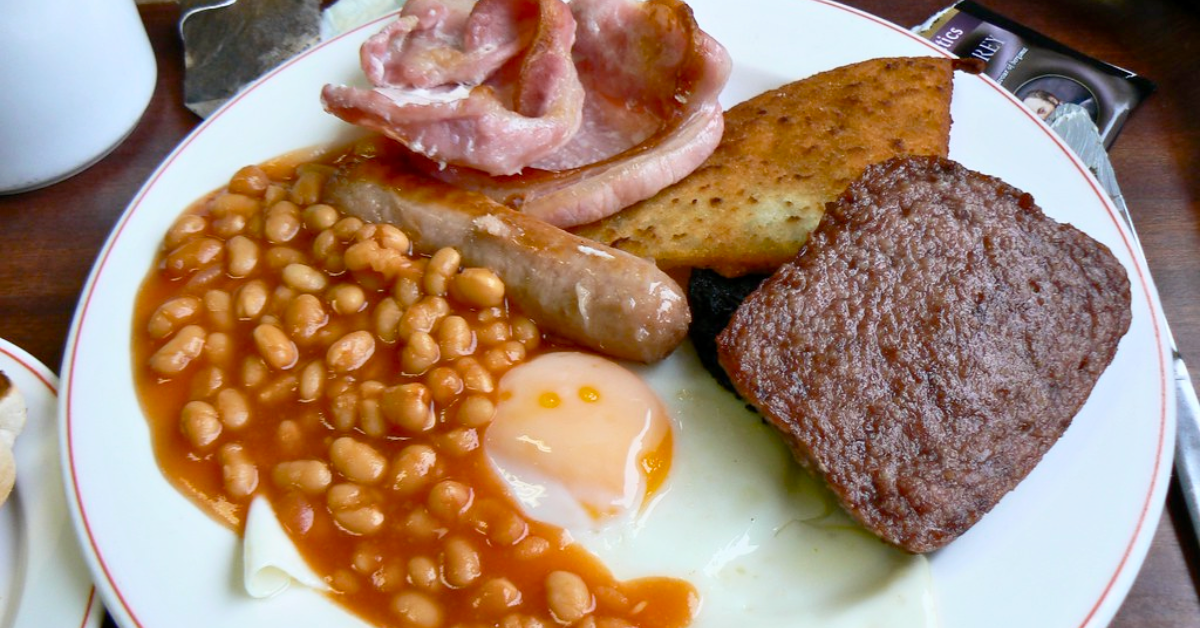 A example of a full Scottish breakfast. This one has a fried egg, saucy beans, a sausage link, a sausage patty, a tattie scone, some thick bacon, and a tiny peek of blood pudding underneath everything.