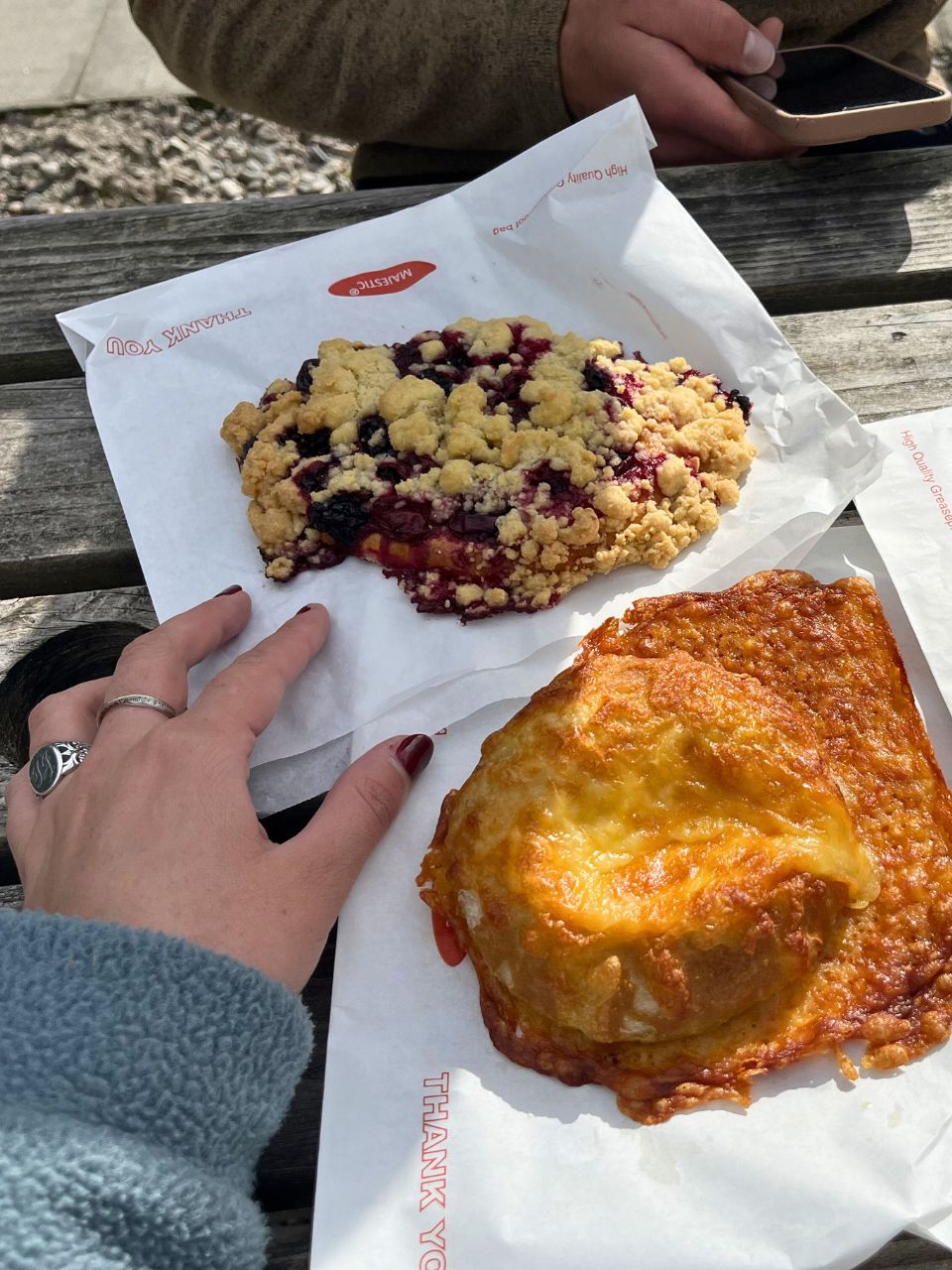 Two baked goods from Manuelas Wee Bakery in Scotland. The bread in front is covered in baked in golden cheese and the item in the back in a Black Forest dessert with blackberry jam and strudel pieces covering it. My hand is on the left for size reference and it looks small compared to both baked goods!