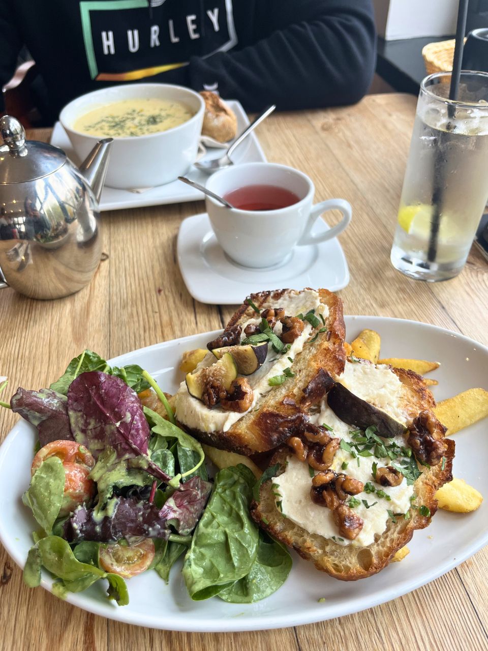 a meal of sourdough break with honey whipped goats cheese, candied walnuts, and fig on top, fries on the side, and a salad drenched in dressing on the left. A hot cup of tea in the middle of the table. A bowl of creamy fish chowder soup in the backgroud.