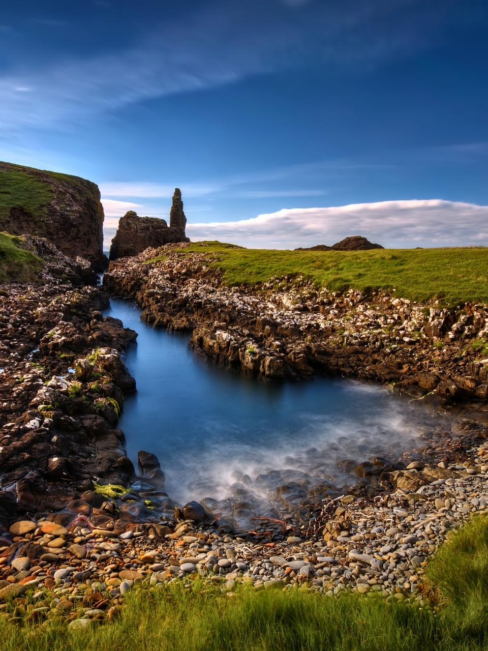 A small inlet of water surrounded by rocks and grass with further rock formations in the background. It is the coastline of Rubha Huish on the Isle of Skye and the ocean is in the distant background. The sky is a vibrant, almost unreal blue shade and the grass is vibrantly green!
