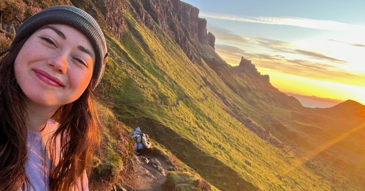 Becca of Oso Osito on the left side of the pic smiling and holding the camera to show the landscape behind her. Rolling green hills, rocky cliffs, the colorful sky, and the sun peeking out on the right hand side. It is sunrise at the Quiraing! Matze can be seen behind her a bit, on the trail, squatting down with his camera to take a picture. The entire photo is bathed in warm, orange light with sunrays shooting across.
