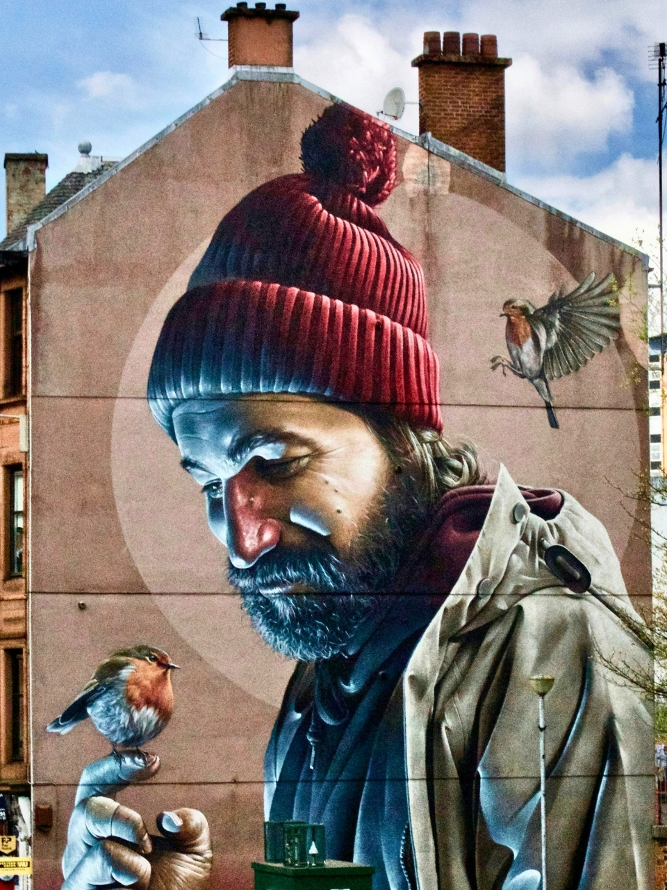 An image of a street art graffiti mural of Saint Mungo in Glasgow City. He wears a red pom pom beanie and has a small bird in his hand.